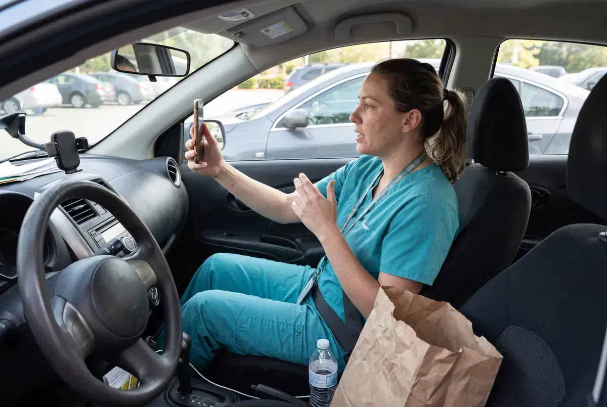 A home care aide in scrubs taking a video interview on her phone in her parked car