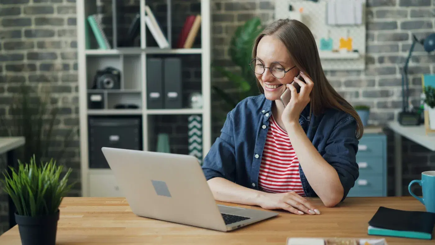 Woman in glasses smiling on a phone call, a laptop open on a wooden desk in a brick-walled office
